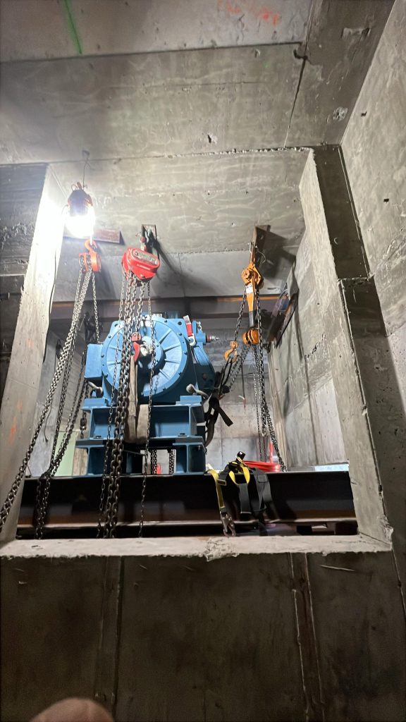 he image shows a geared traction machine for an elevator, located in a machine room. This type of machine is used in traction elevators to lift and lower the elevator car by winding and unwinding steel cables on a large wheel called a sheave. The multiple chain hoists and rigging equipment visible are used for the installation, removal, or maintenance of heavy components in the elevator shaft